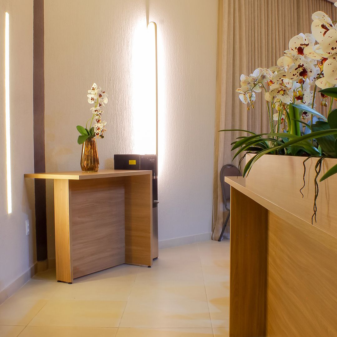 Quarto de hotel com cama, TV, mesa e cadeiras. Detalhes em madeira, cortinas brancas e uma luminária retangular no teto.