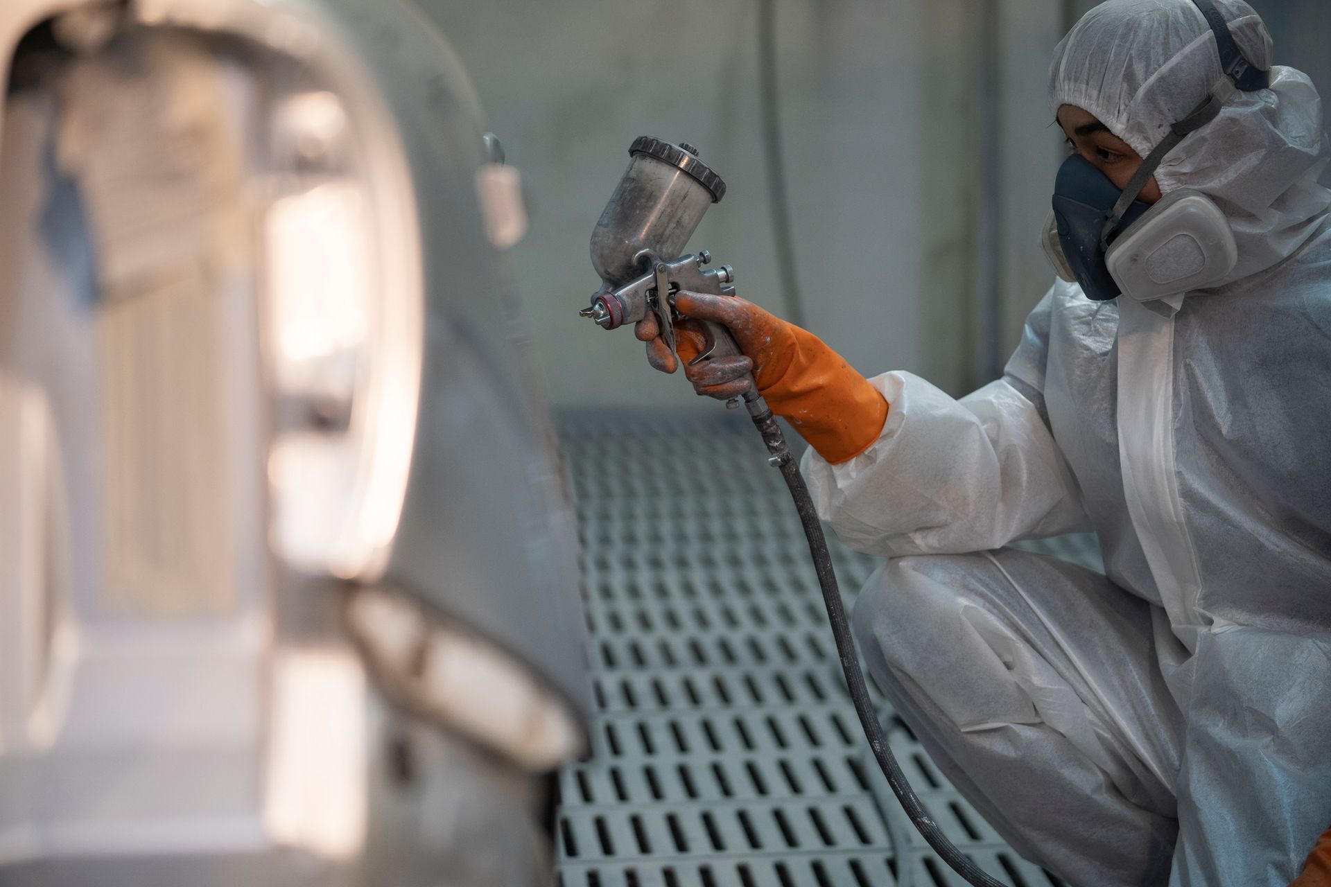 Una persona con tuta protettiva e respiratore vernicia a spruzzo un componente di un'auto in un'officina; indossando guanti arancioni, è inginocchiata vicino a una grata metallica.