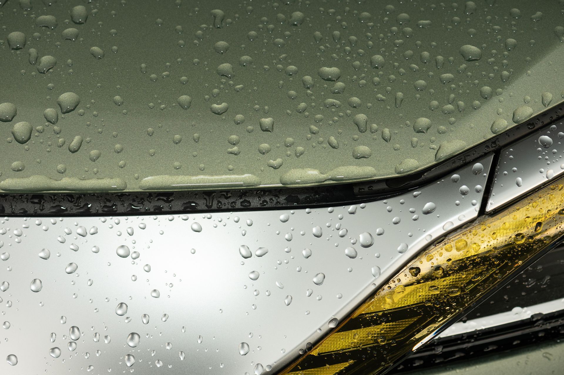 Gocce di pioggia sul cofano e sul faro di un'auto color argento metallizzato. La vernice ha una tonalità verde chiaro.