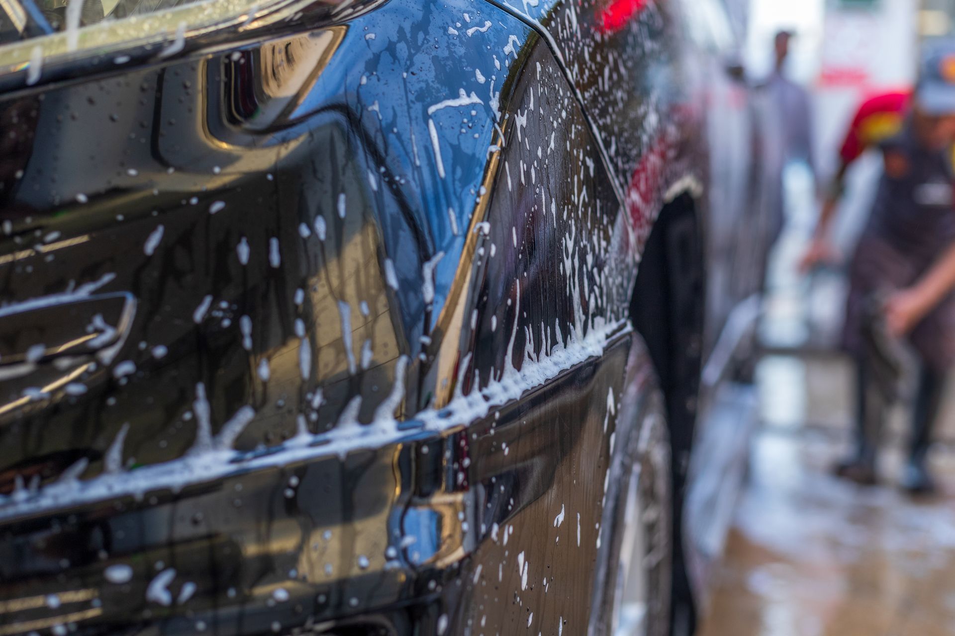 Un'auto nera viene lavata in un autolavaggio, ricoperta di schiuma di sapone. Sullo sfondo c'è una persona.