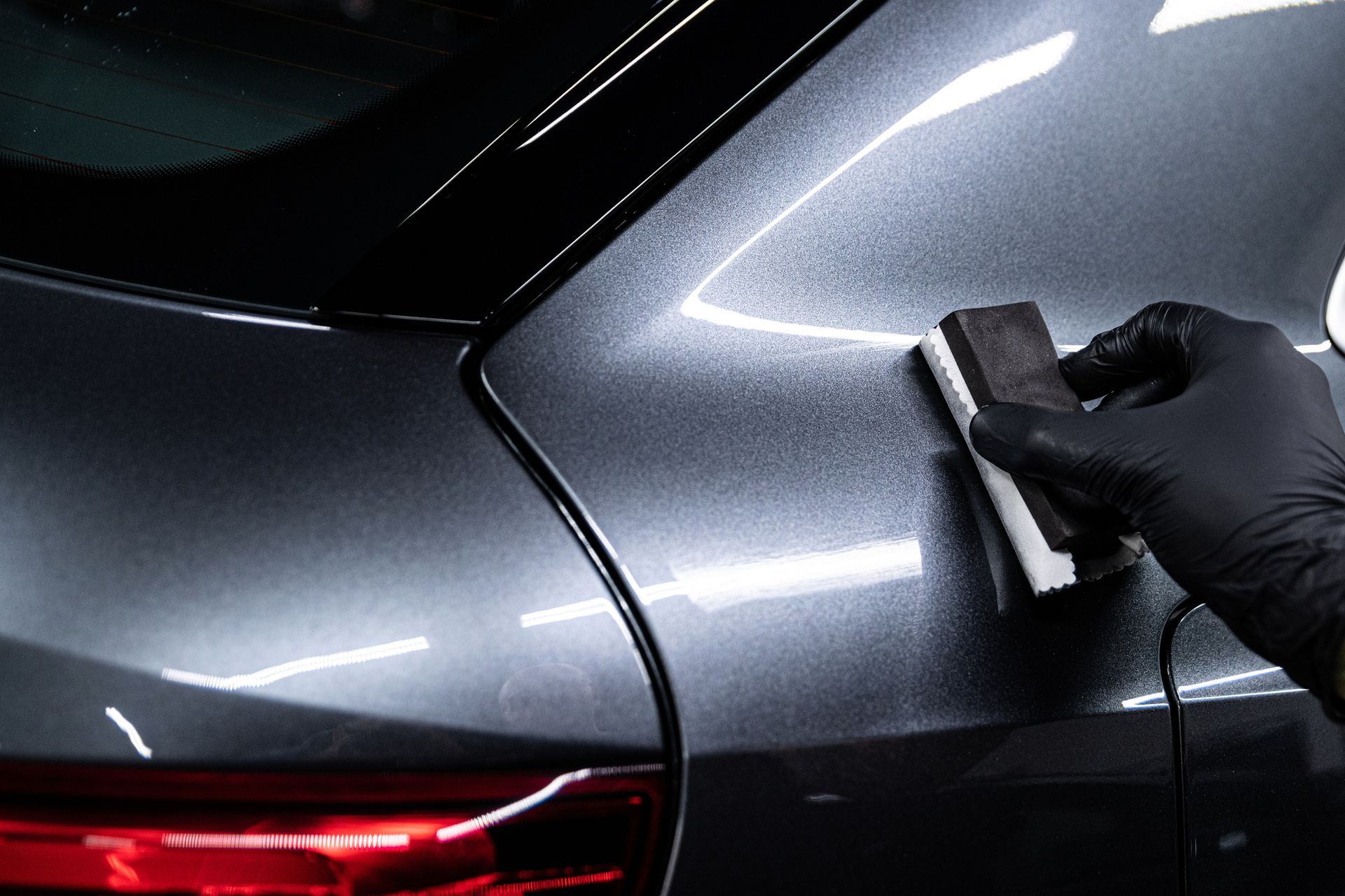 Mano guantata che applica un rivestimento ceramico su un pannello grigio scuro di un'auto. La superficie riflette la luce e l'applicatore è in uso.