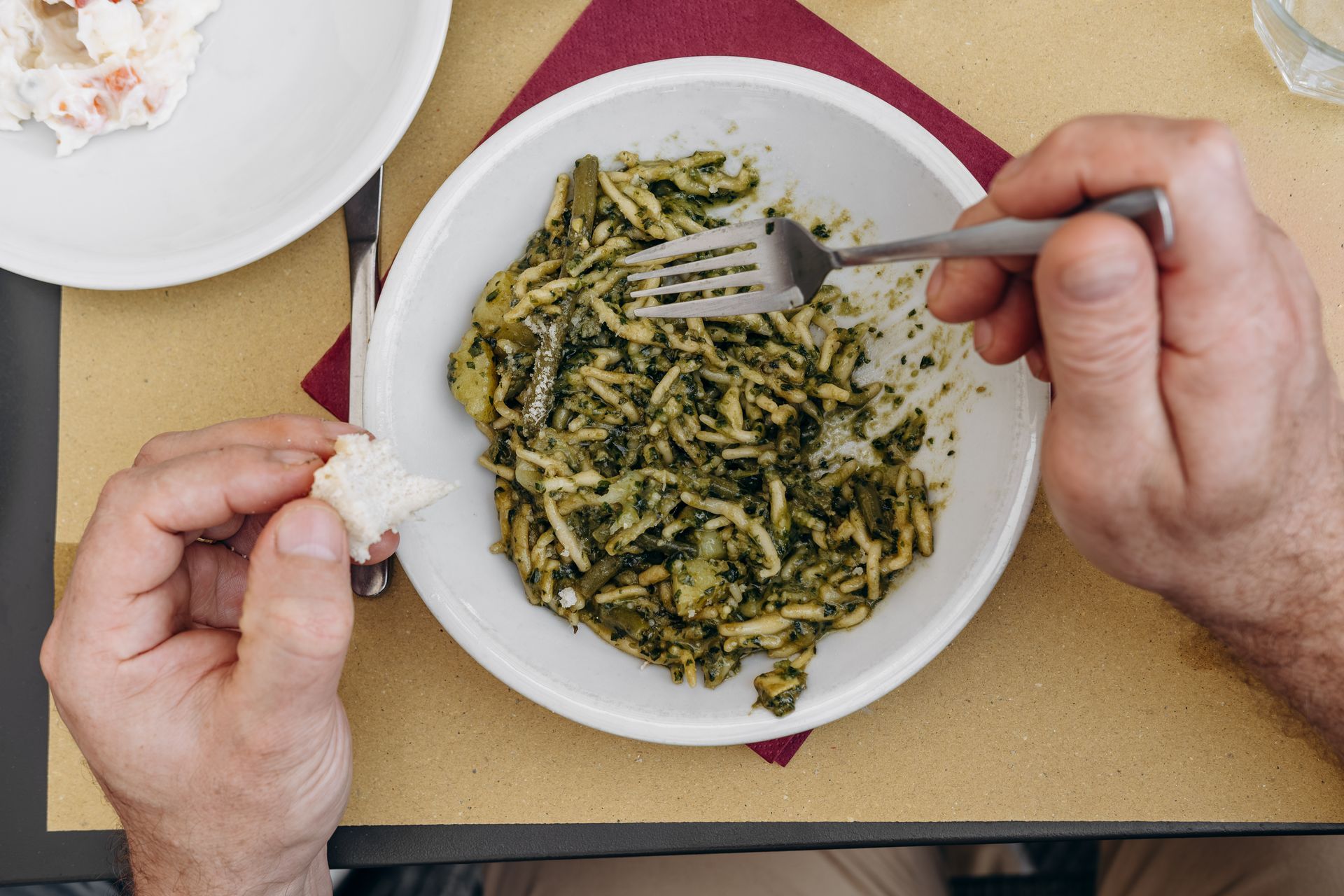 Persona che mangia pasta al pesto da una ciotola bianca, usando una forchetta e un pezzo di pane.
