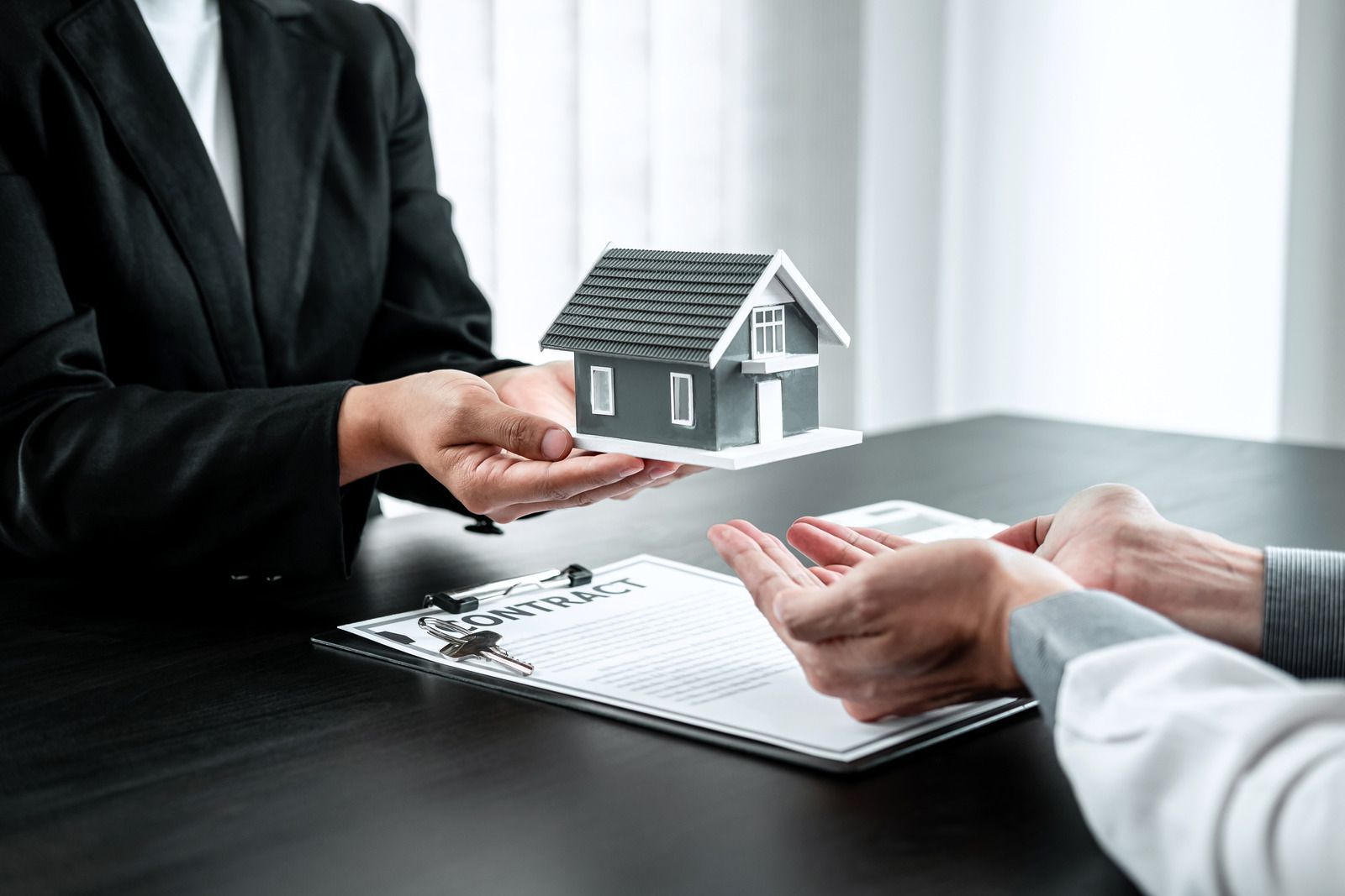 Real estate agent handing a miniature house to a client at a desk, paperwork present.