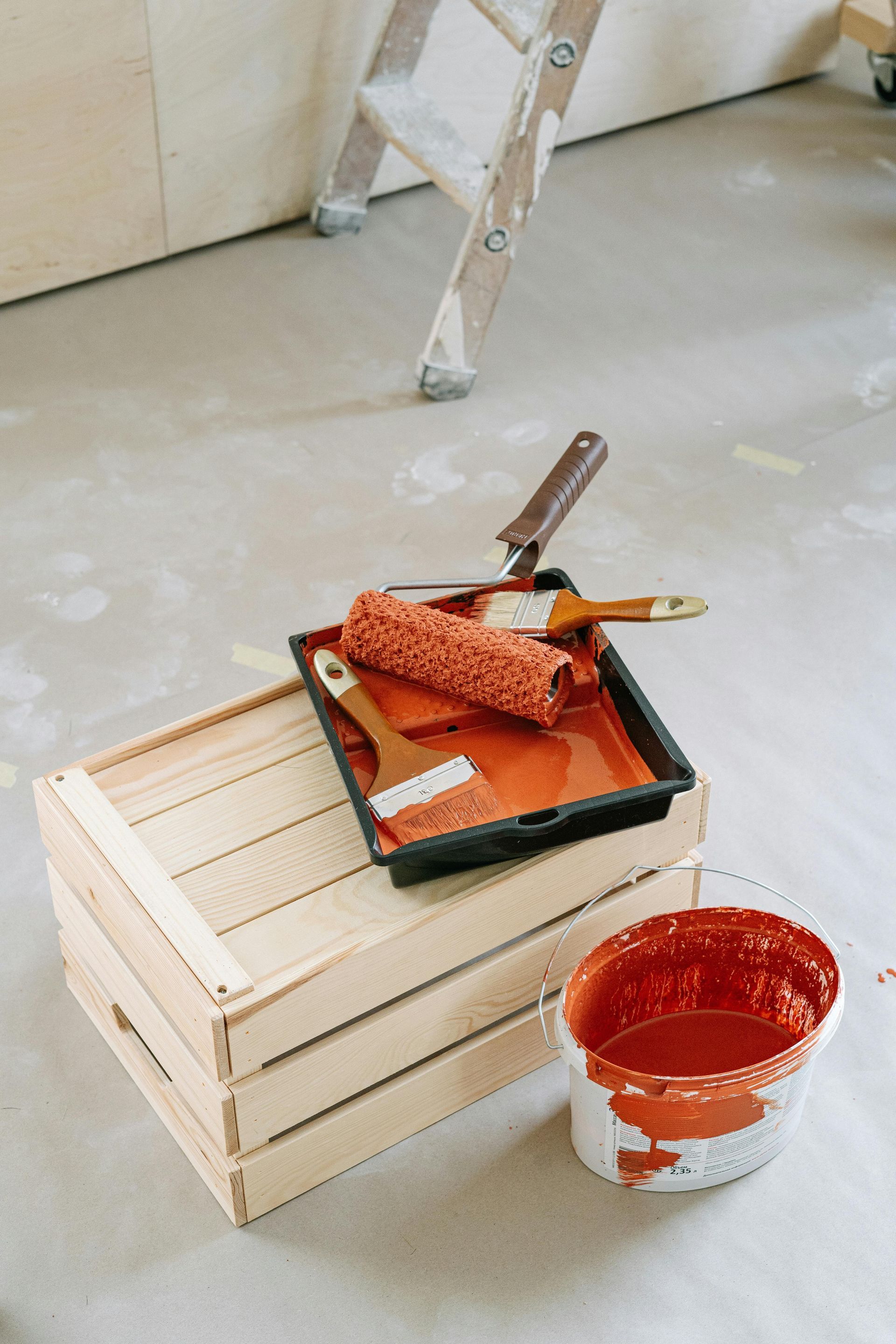 Paint tray with orange paint, roller, brush, and bucket on a floor beside a wooden crate and step ladder