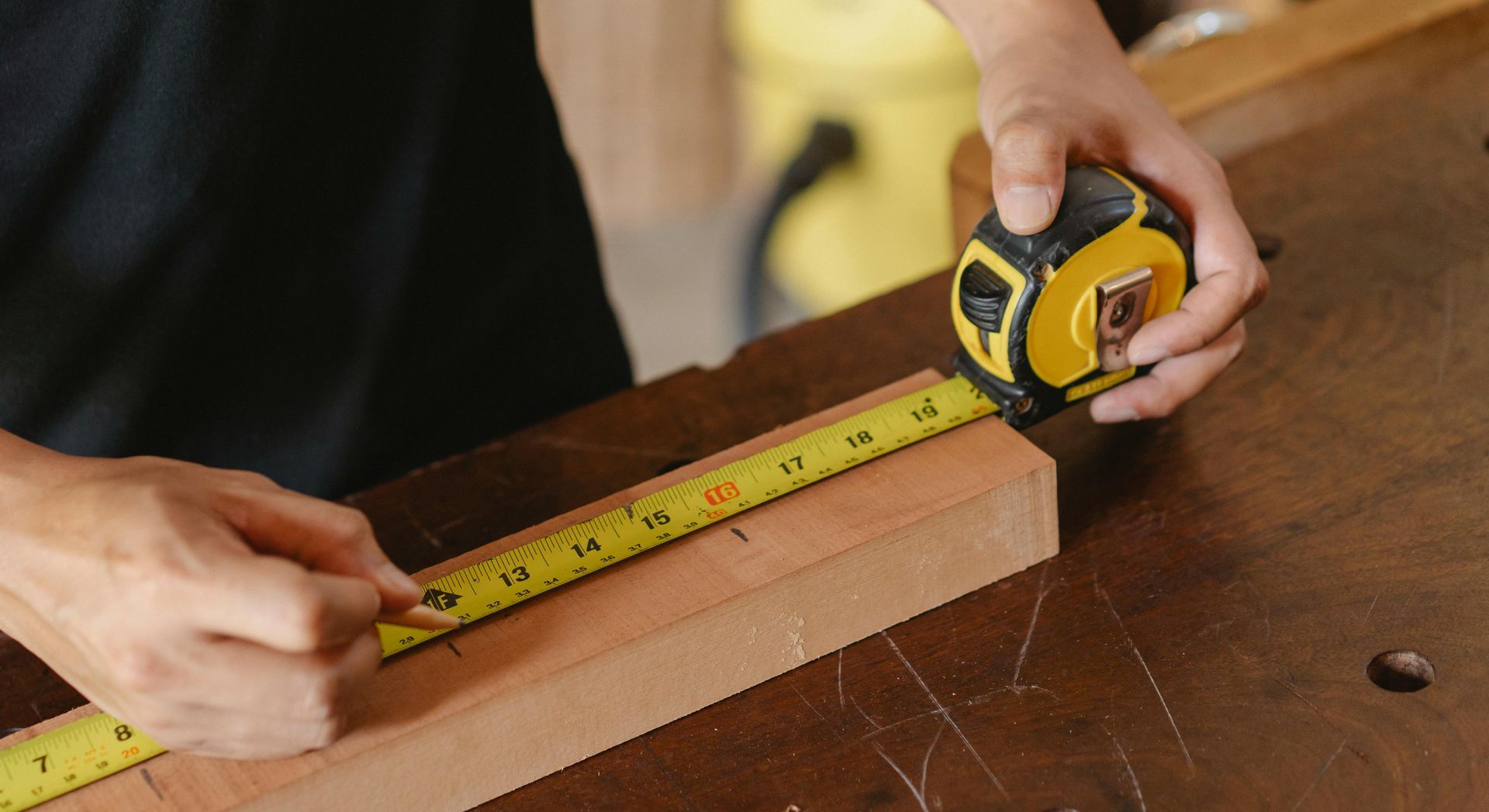 Hands measuring a wooden board with a yellow tape measure on a workbench.