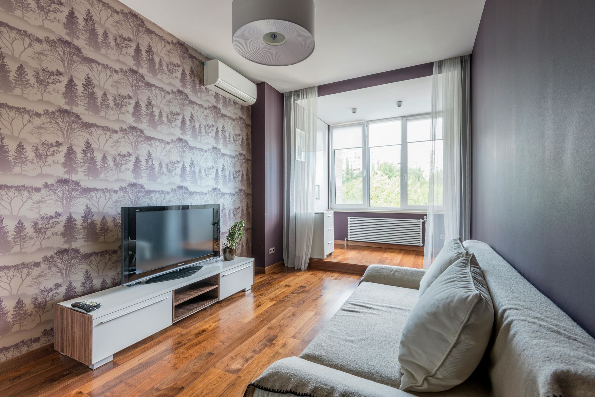 Modern living room with gray sofa, TV on white stand, wood floor, and large window seating area