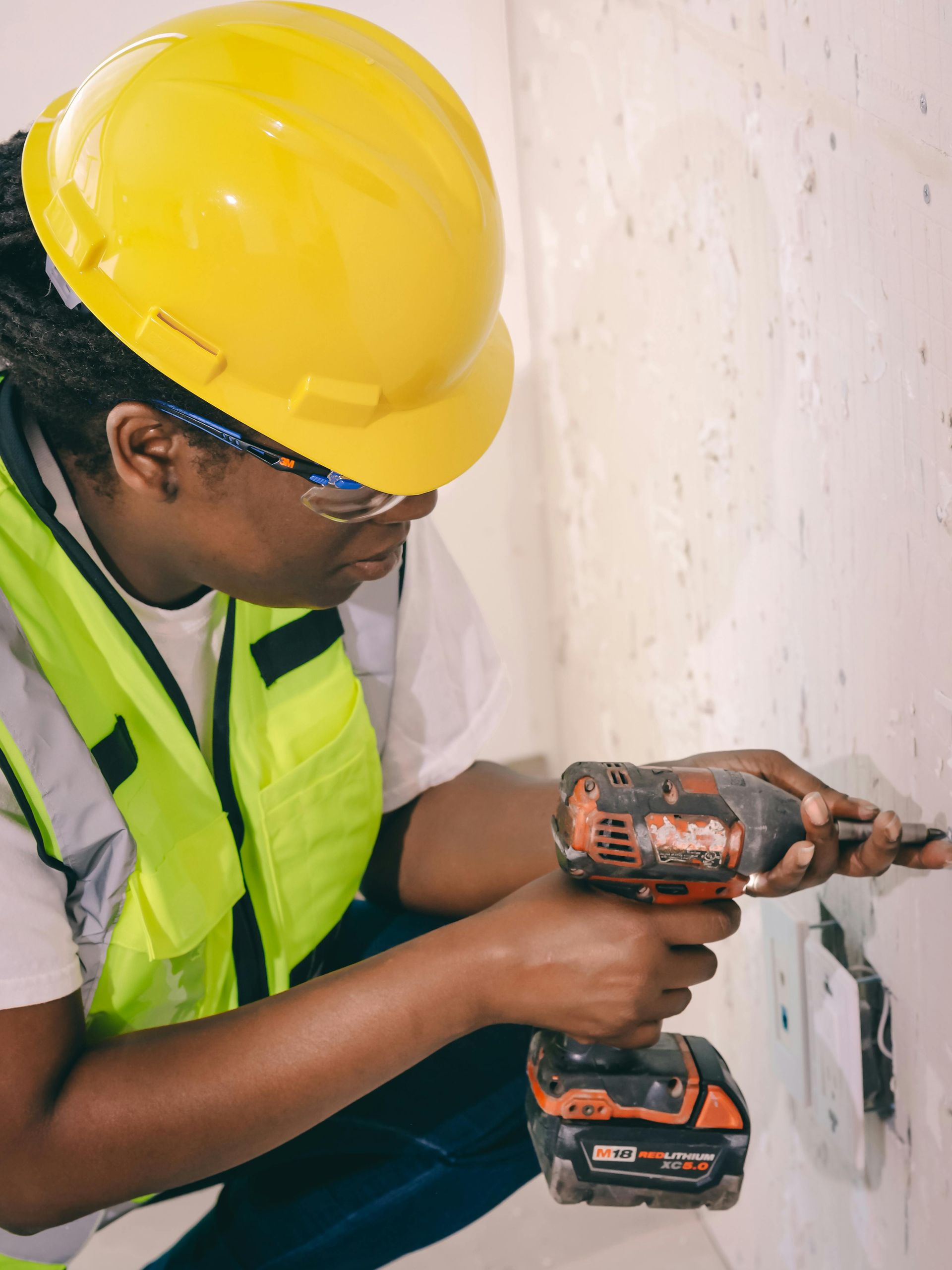 Worker in yellow hard hat drills into a wall near an electrical outlet.