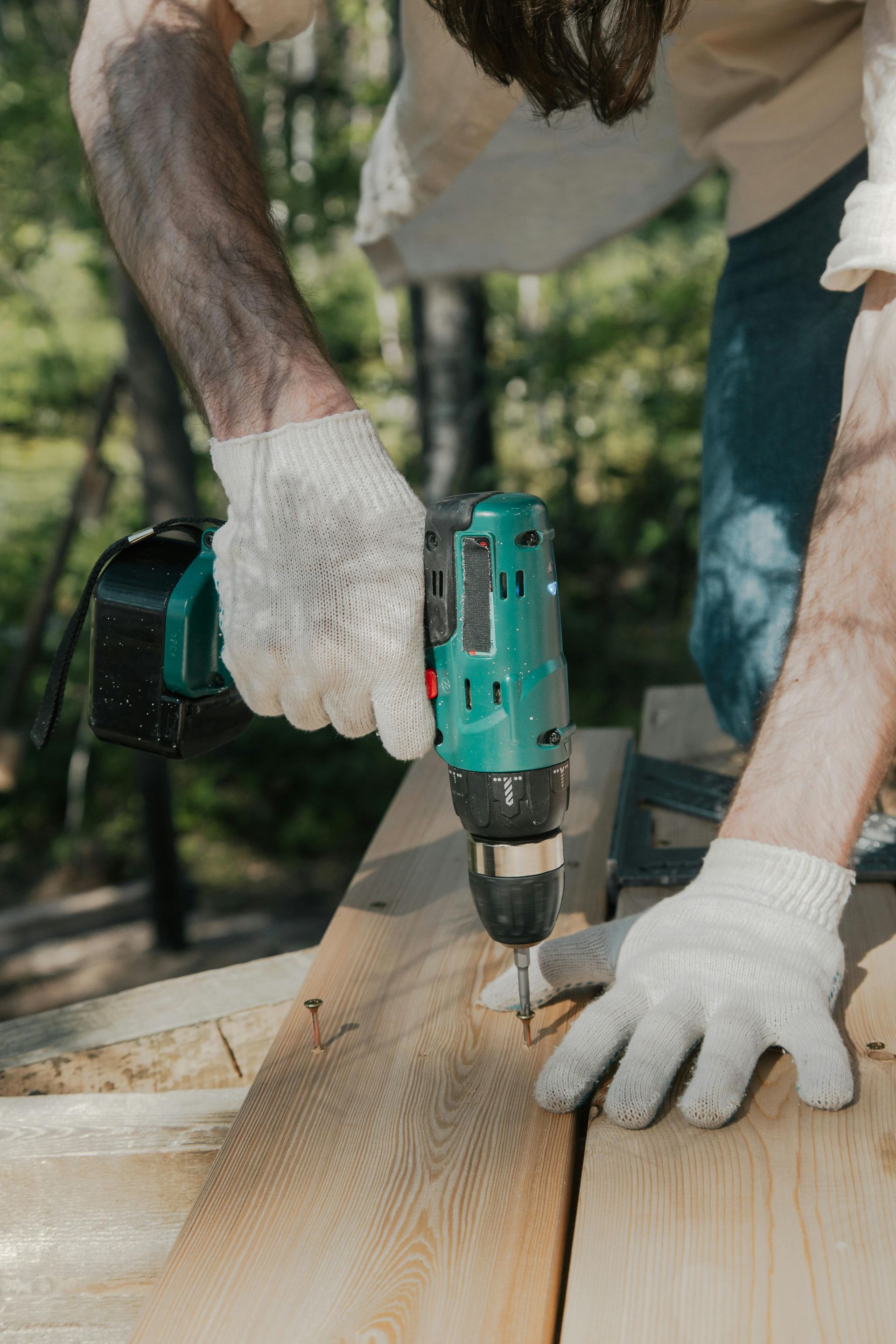 Person drilling a wooden plank outdoors with a teal power drill and white gloves