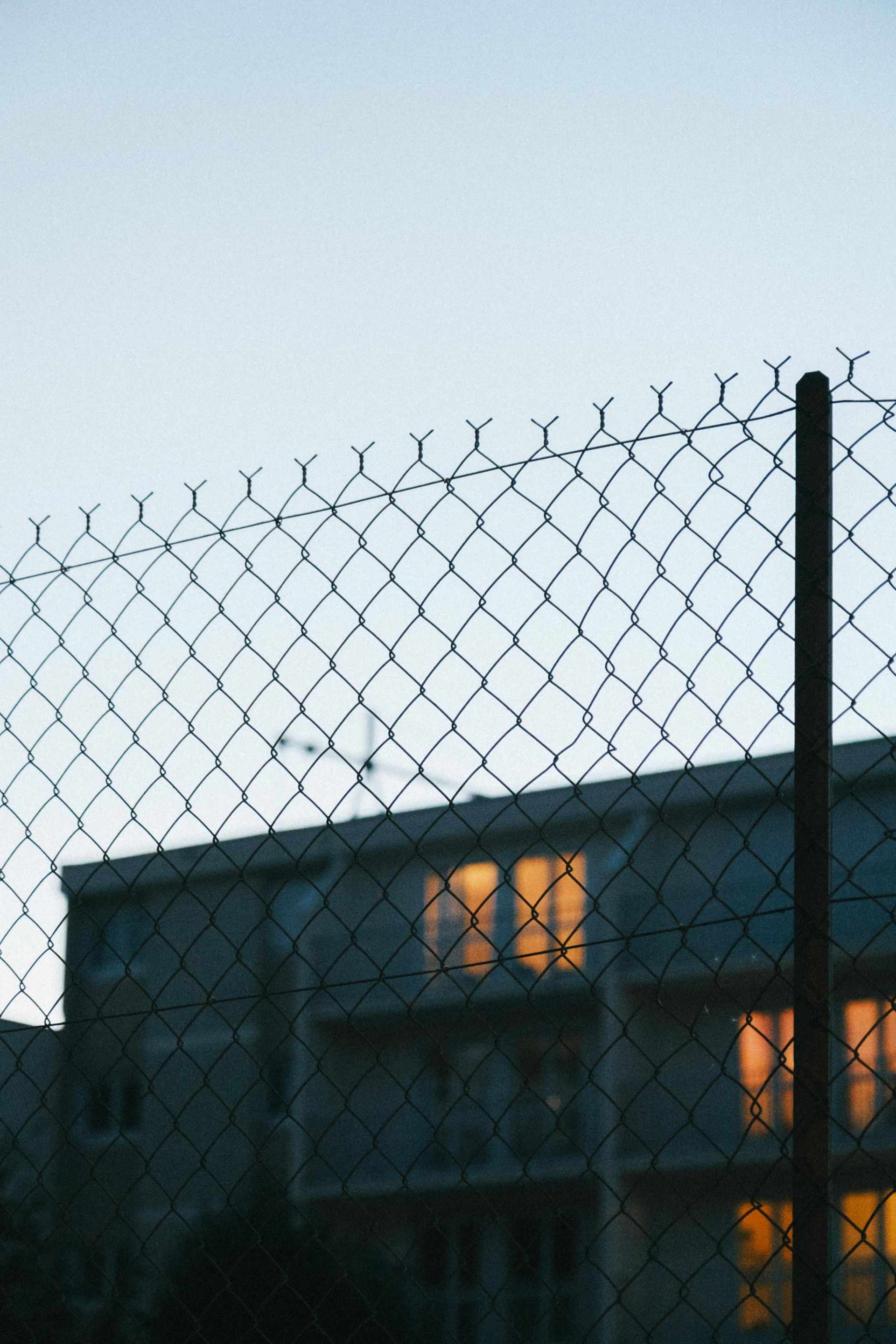 Chain-link fence in front of an apartment building at dusk, with warm lights glowing in the windows.