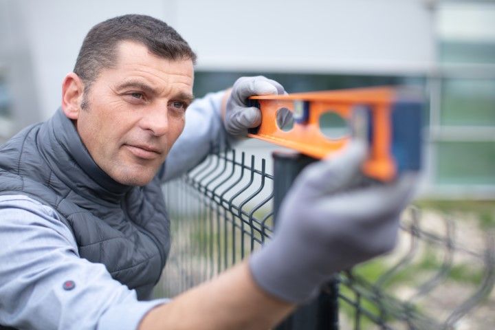 Man using a level to check fence post alignment outdoors, wearing gloves and vest.
