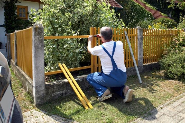 Man in blue overalls kneels, installing a yellow wooden fence section between concrete posts, outdoors on a sunny day.