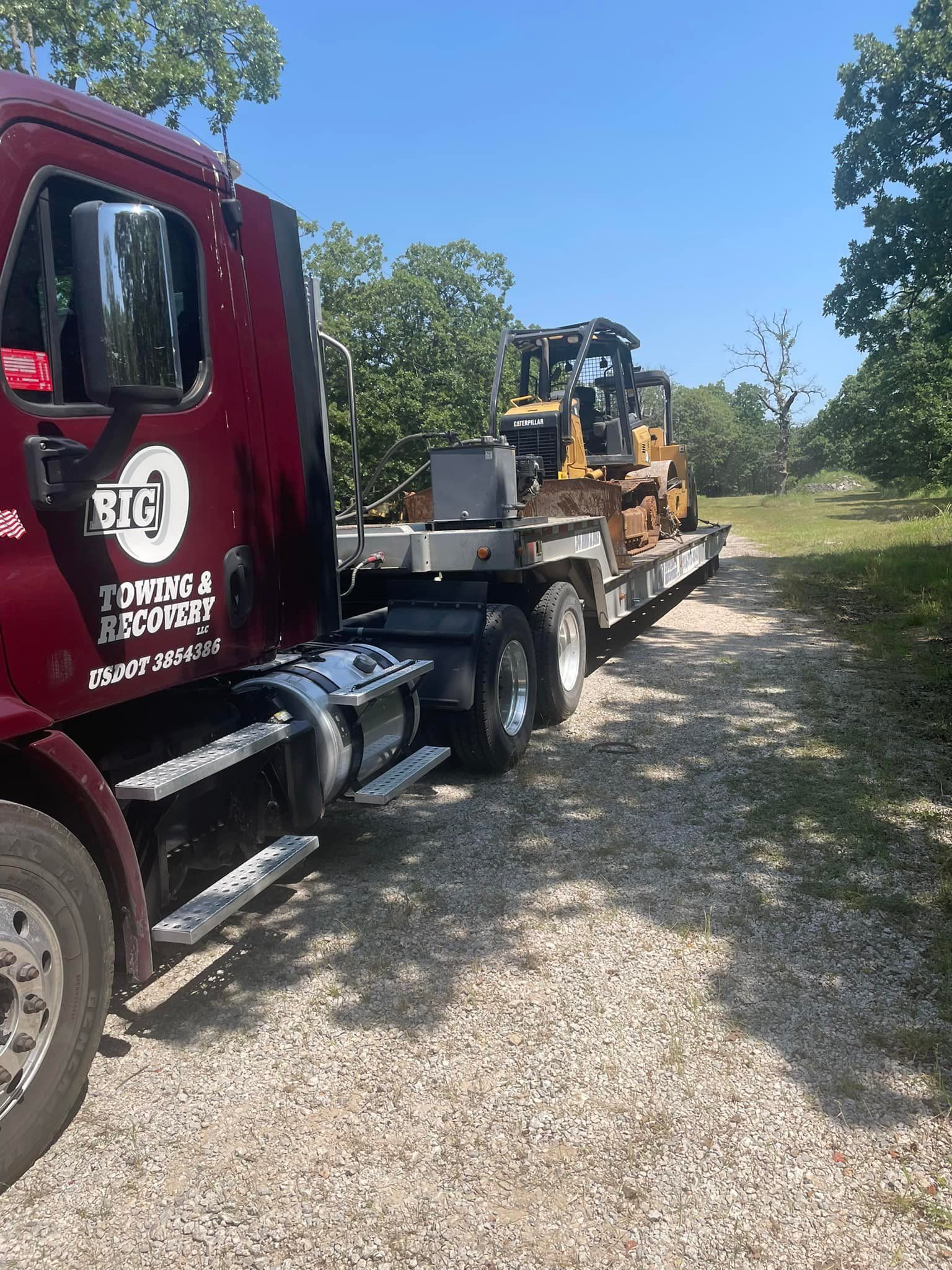 Maroon semi-truck towing a yellow bulldozer on a trailer along a gravel path.