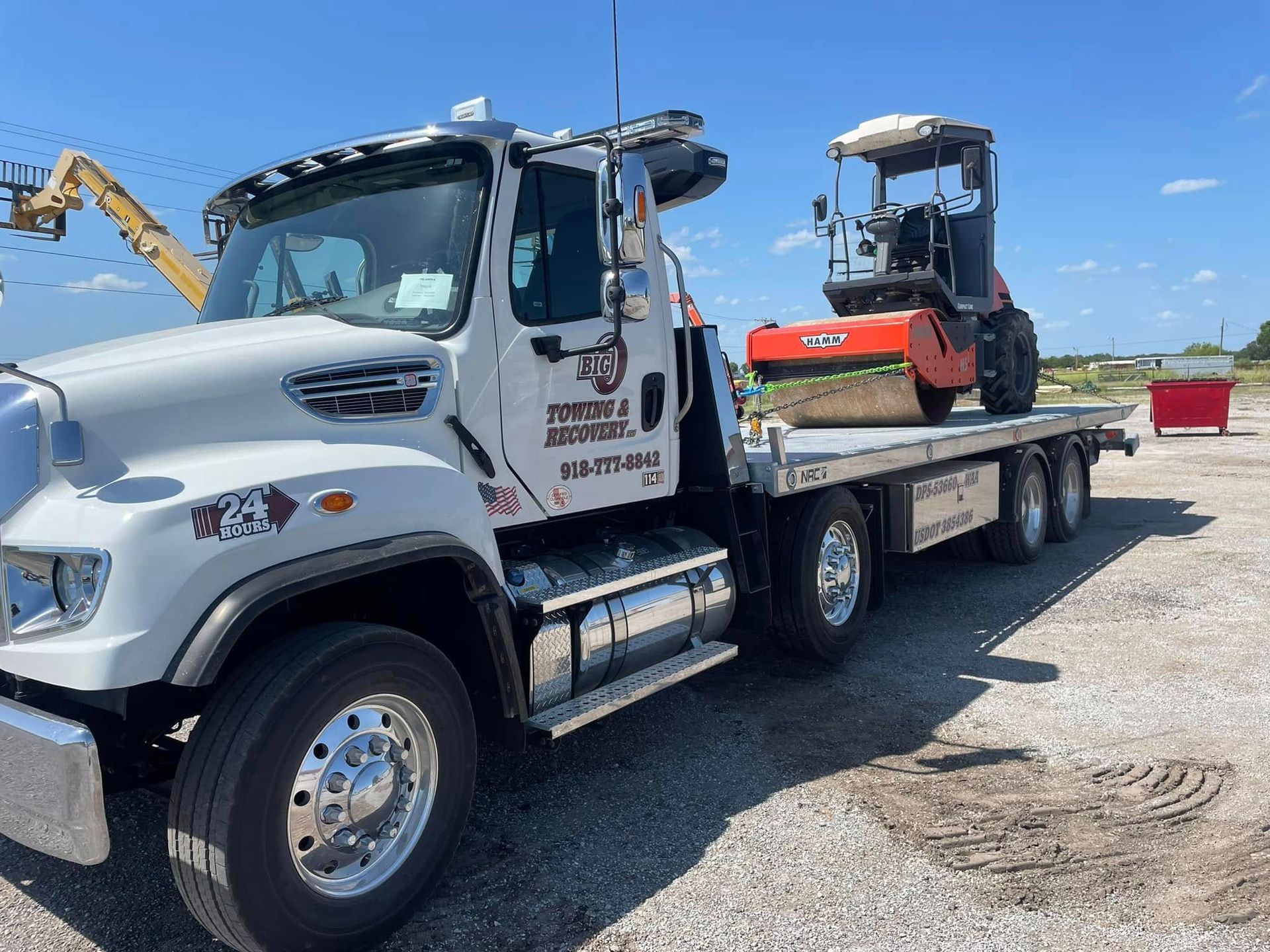 White flatbed truck transporting a small road roller on a sunny day.