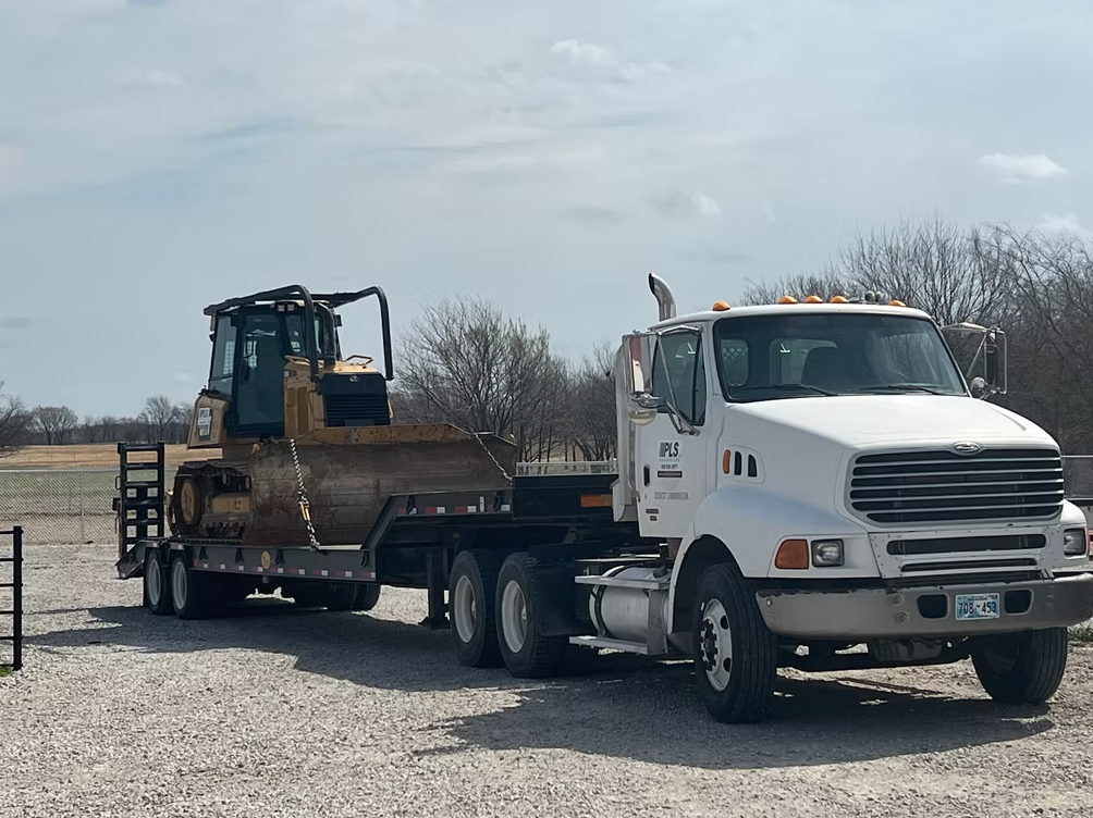 Truck transporting a bulldozer on a flatbed trailer. On gravel, outdoors. Bright, daytime.