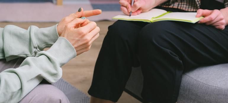 A woman is sitting on a couch talking to a man who is writing in a notebook.