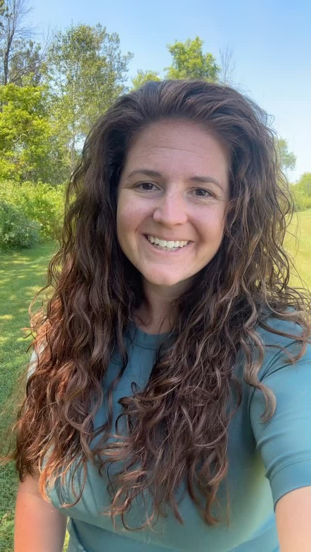 Woman with curly brown hair smiles outdoors, wearing teal shirt, with trees and blue sky in background.