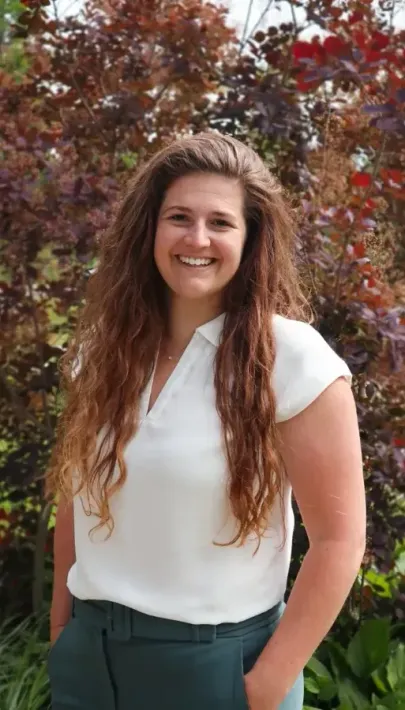 Woman with curly brown hair smiles, wearing a white top and green pants, standing in front of a tree.