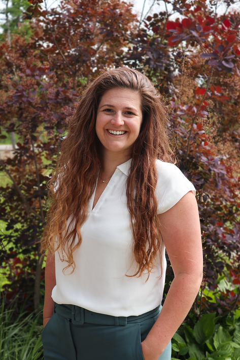 Woman with curly brown hair smiles, wearing a white top and green pants, standing in front of a tree.