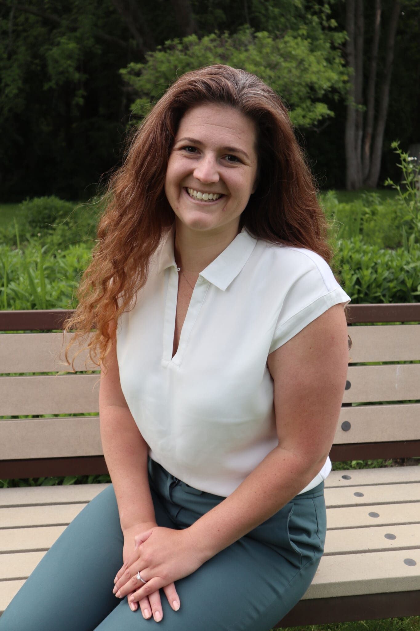 Woman with curly red hair, sitting on a bench, wearing a white shirt and teal pants, smiling.