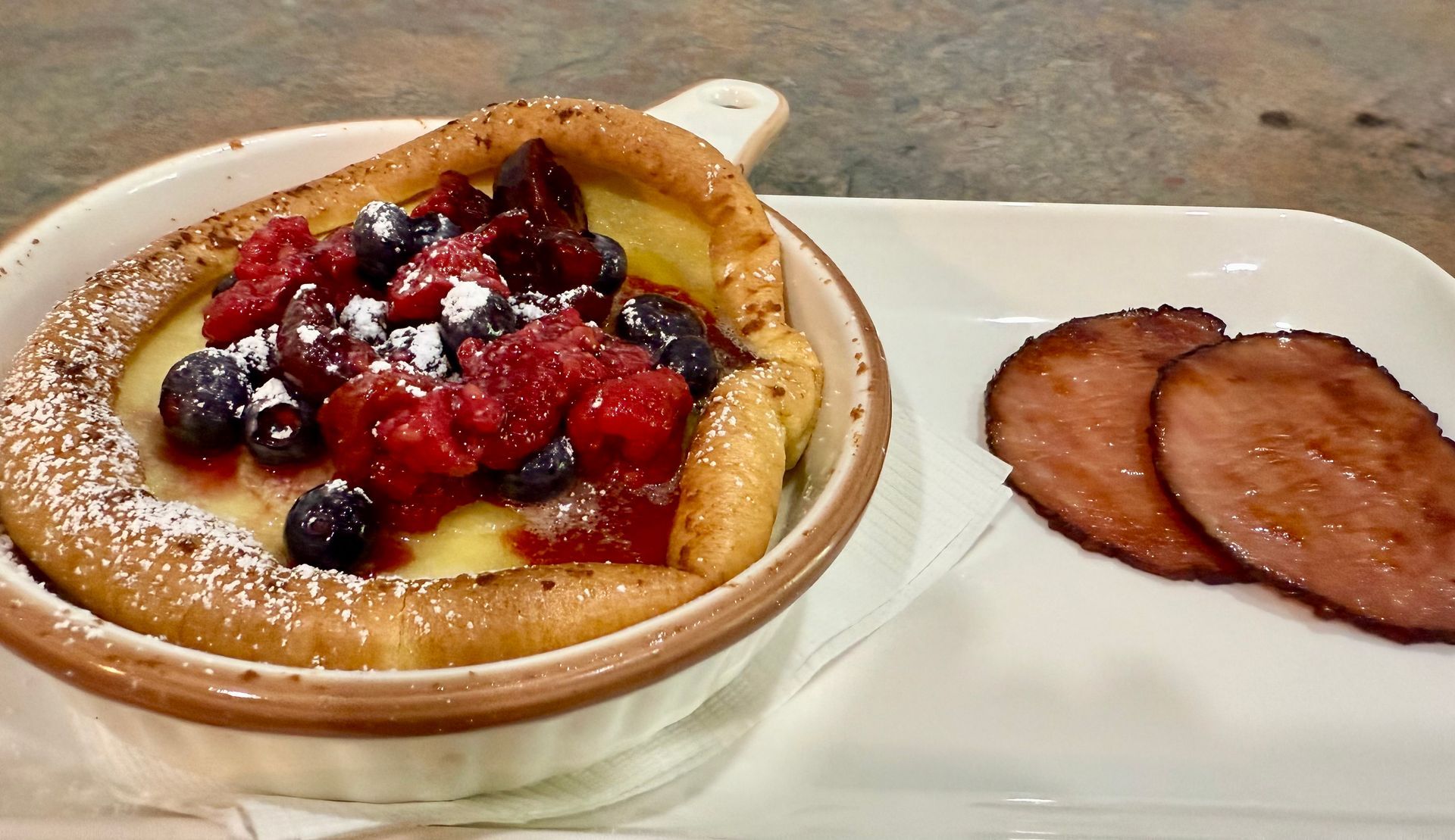 A bowl of food with berries and powdered sugar next to a plate of ham