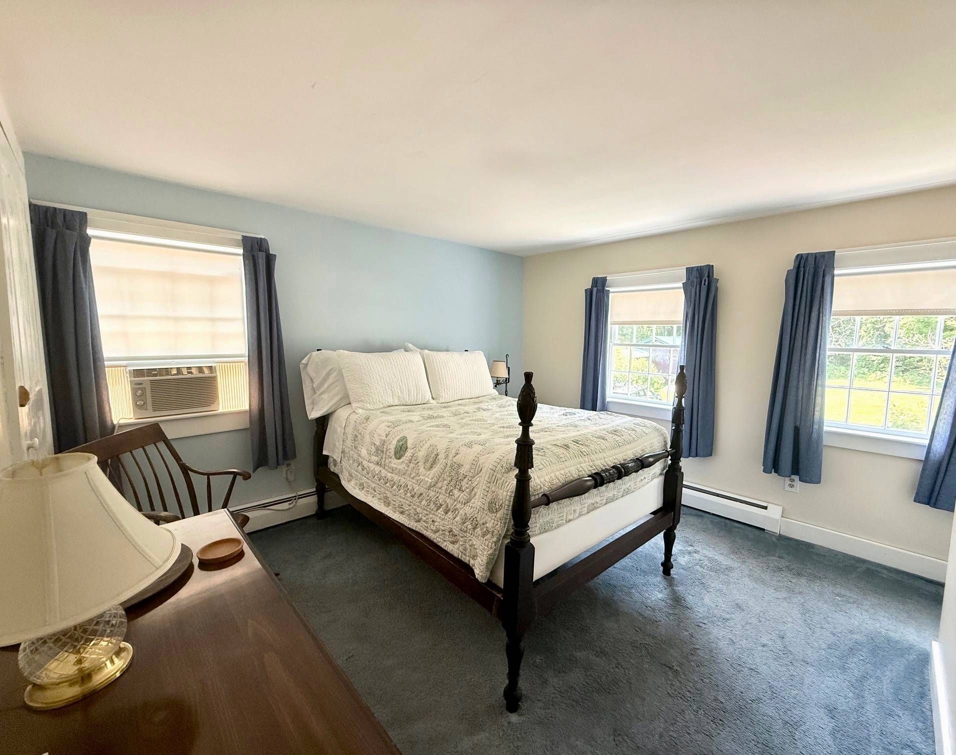 A bedroom with a bed , dresser , chair , lamp and windows.