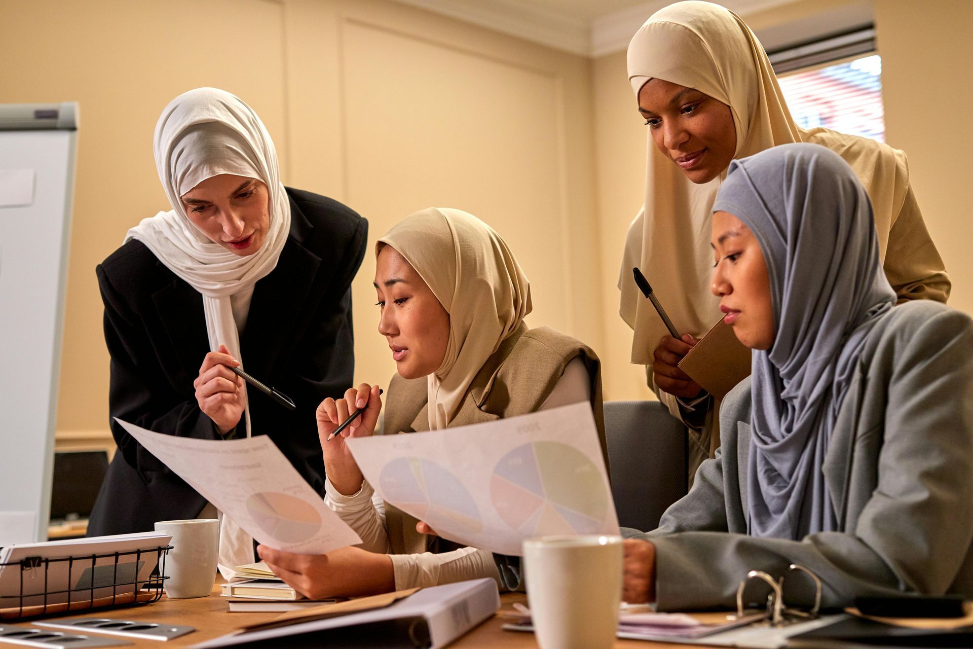 Four women in hijabs and professional attire collaborating in an office, reviewing charts and discussing work.