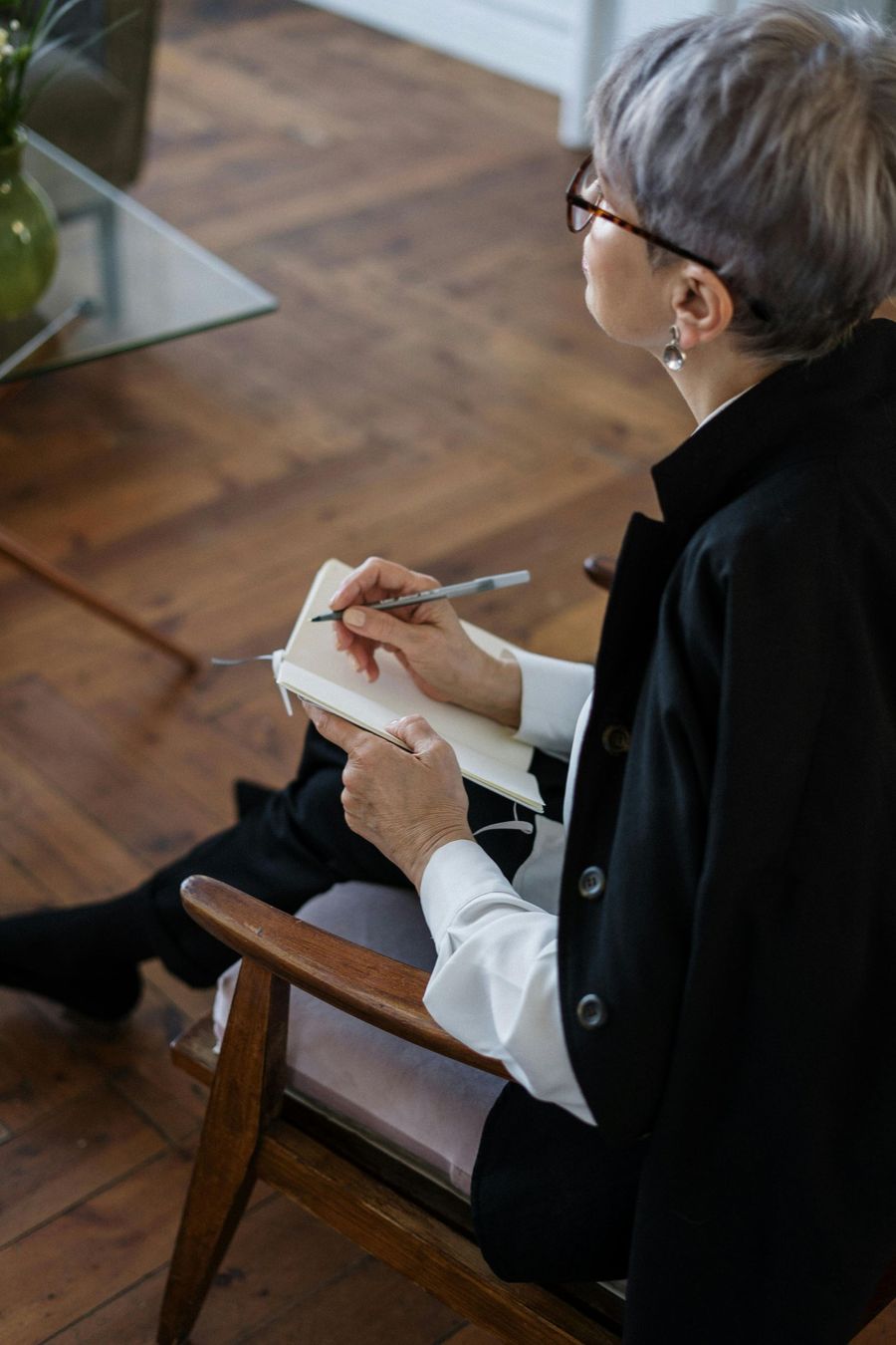 A person in a black jacket and white shirt sits in a wooden chair, writing in a notebook while looking away.