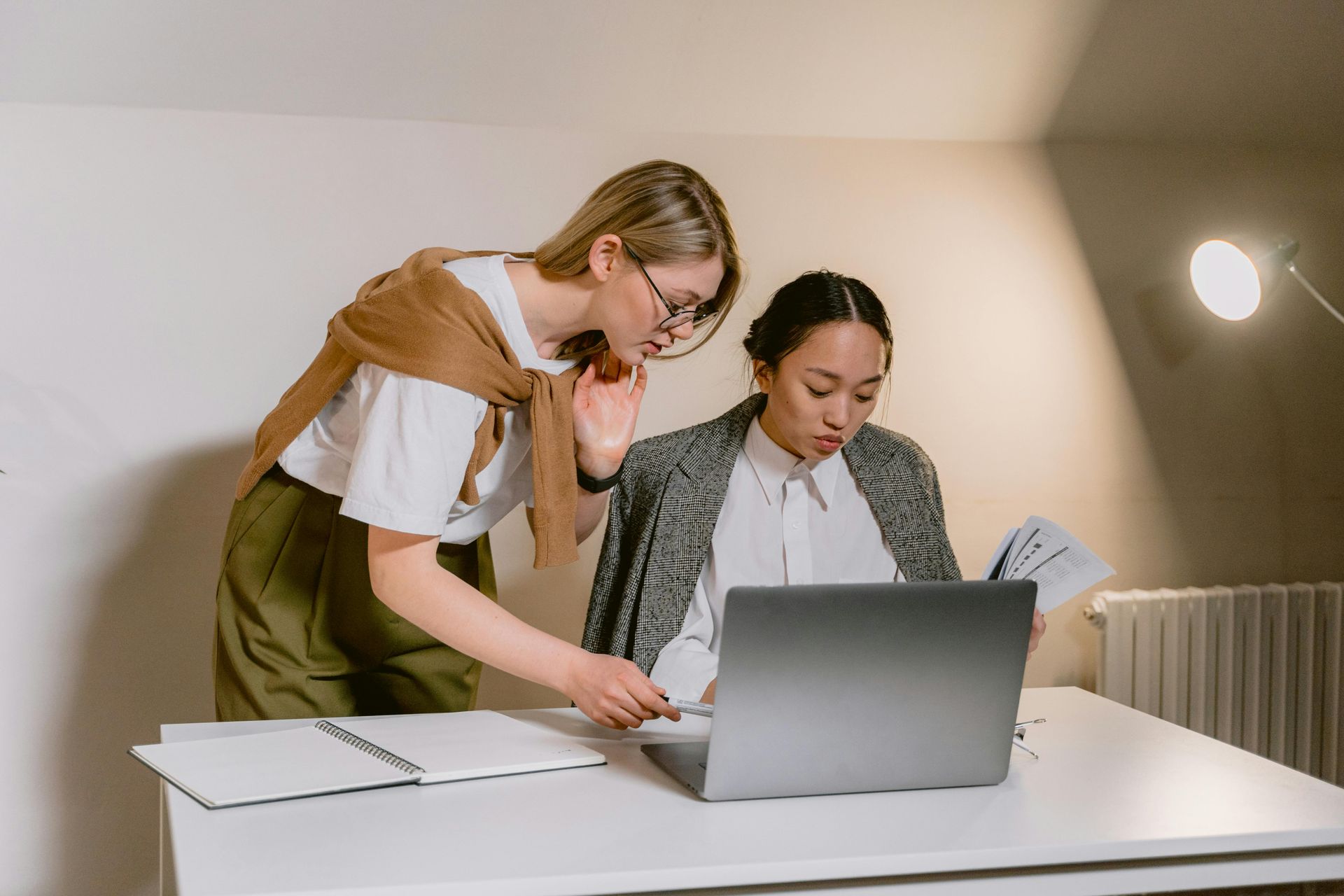 Two people collaborate at a desk with a laptop and open notebook in a brightly lit room.