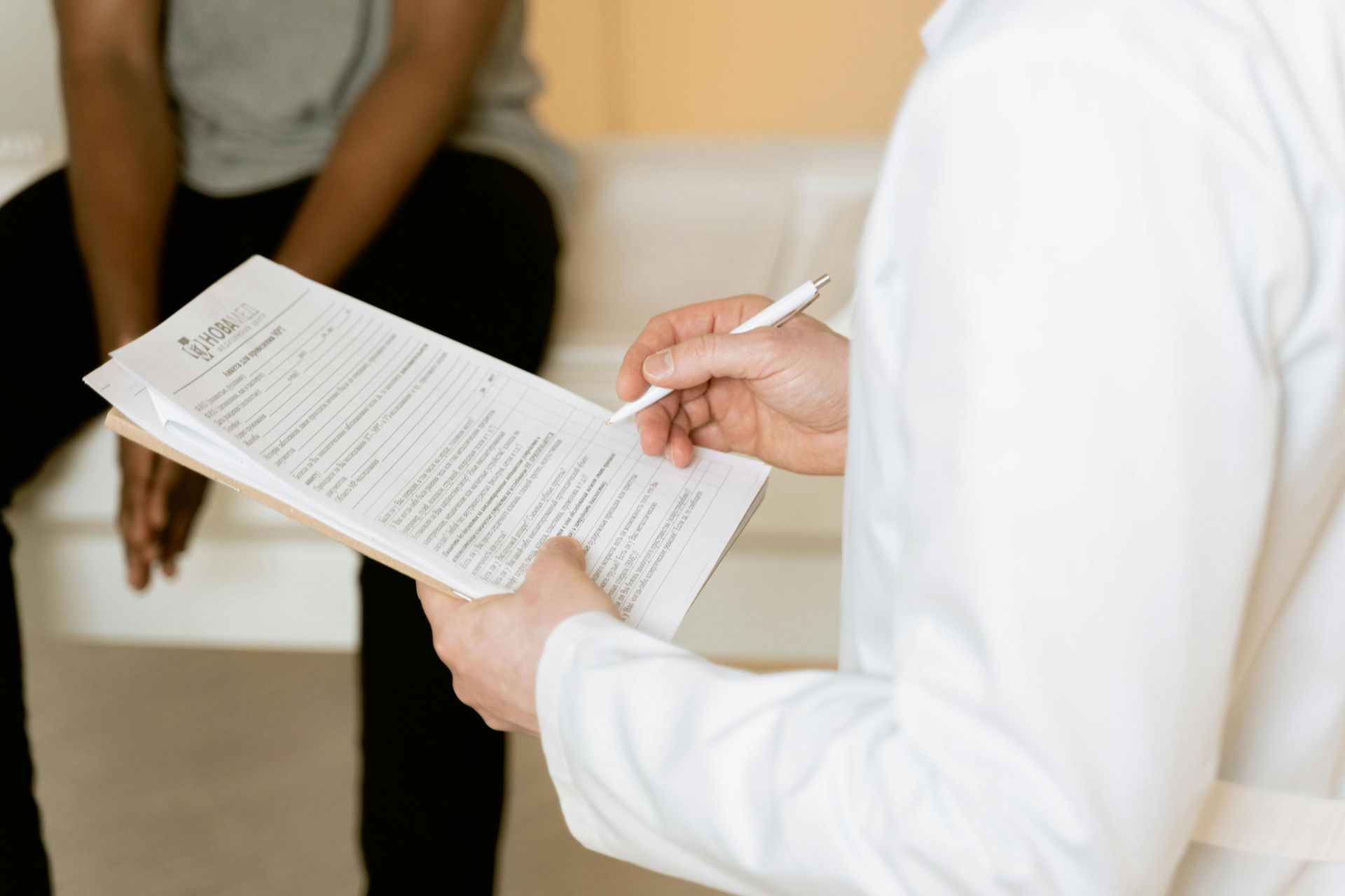 A healthcare professional in a white coat holds a clipboard and pen while consulting with a patient in an office.