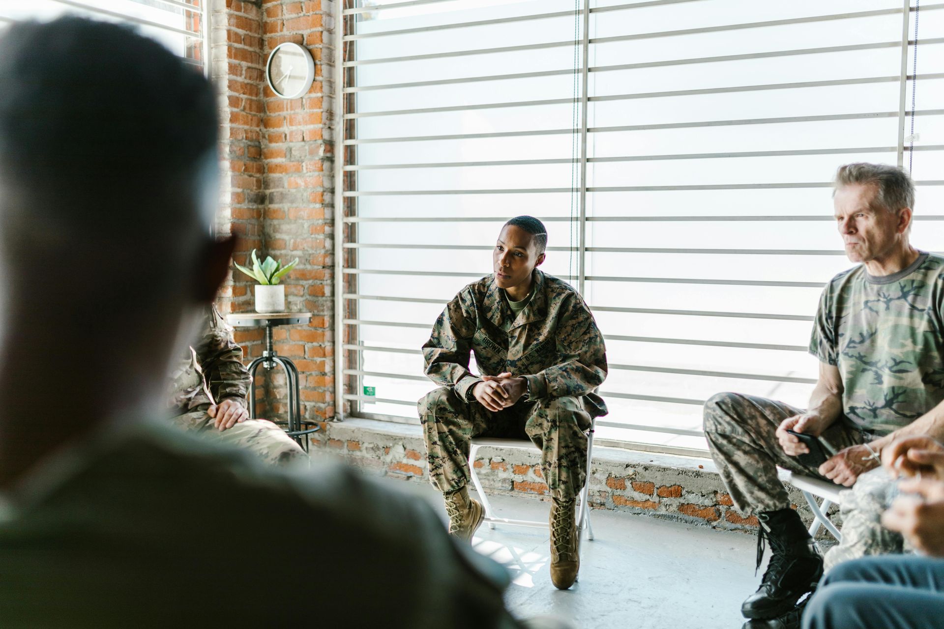 People in military camouflage uniforms sitting in a circle during a group meeting in a room with large windows.