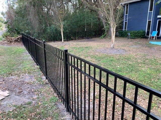 Black metal fence in a yard, surrounded by grass and trees, with a blue house in the background.