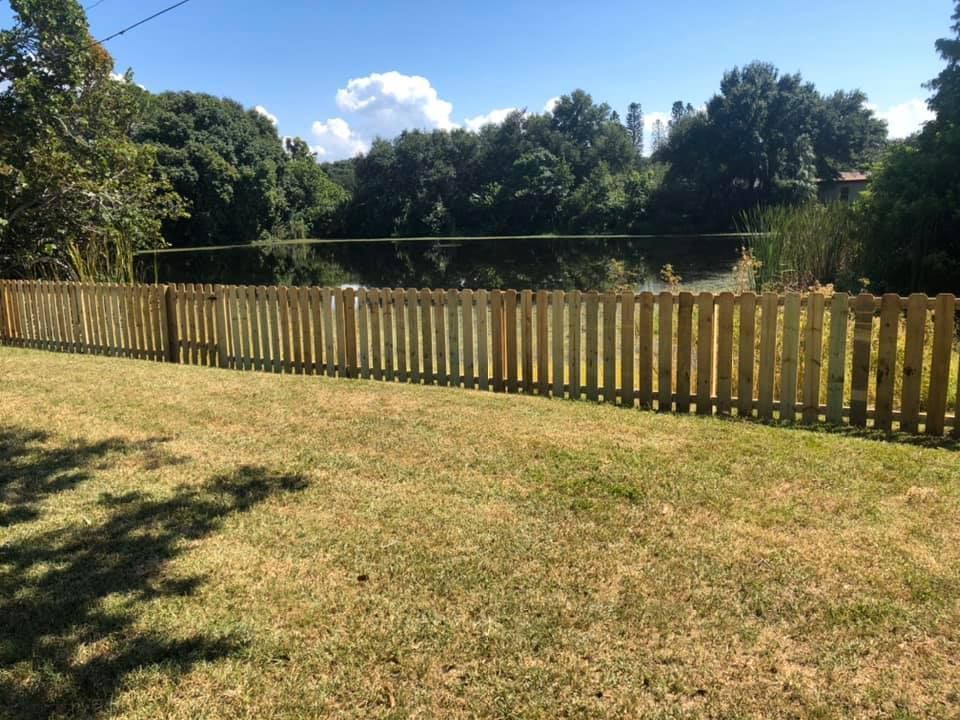 A wooden fence runs along a grassy area with a lake and trees in the background on a sunny day.