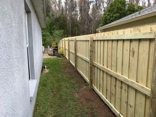Wooden fence along a house's side, with a grassy yard in between. Trees in the background.