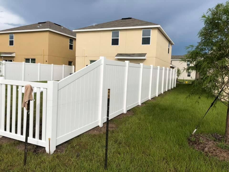 White vinyl fence surrounds a backyard with two beige houses. Overcast sky, green grass.