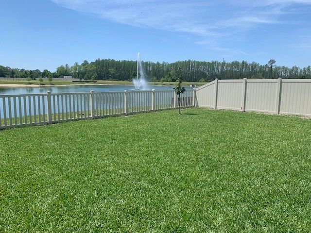 Green lawn fenced next to a pond with a fountain under a blue sky.