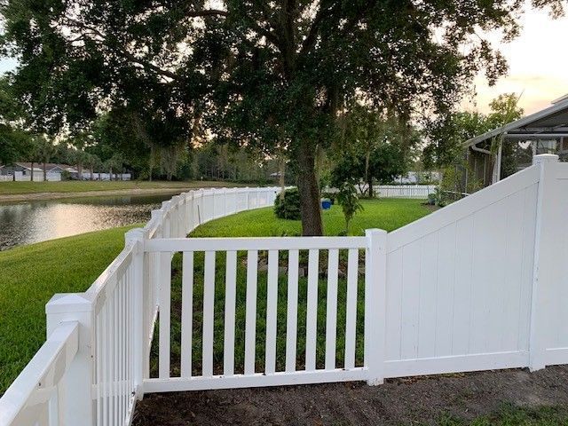 White fence with gate along a lake. Green grass, trees, and houses in the background.