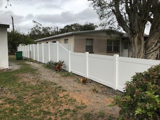 White vinyl fence bordering a one-story brick house with a large tree and overcast sky.
