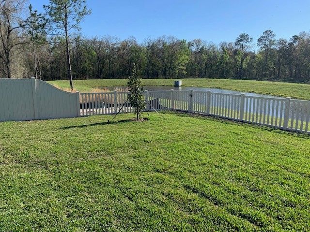 Grassy backyard with white fence, a small tree, and a pond on a sunny day.
