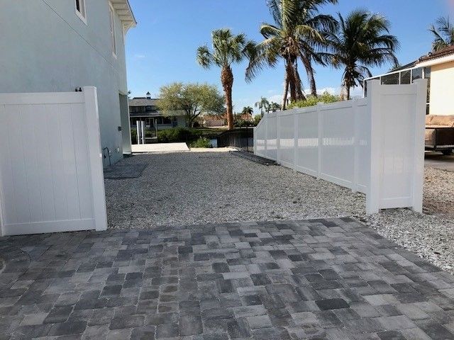 Driveway with white fences and gravel. Palm trees and blue sky in background.