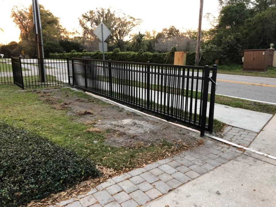 Black metal fence bordering a sidewalk and roadside, with green grass and trees in the background.
