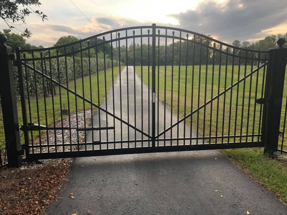 Black metal driveway gate leading to a grassy field, set against a cloudy sky.