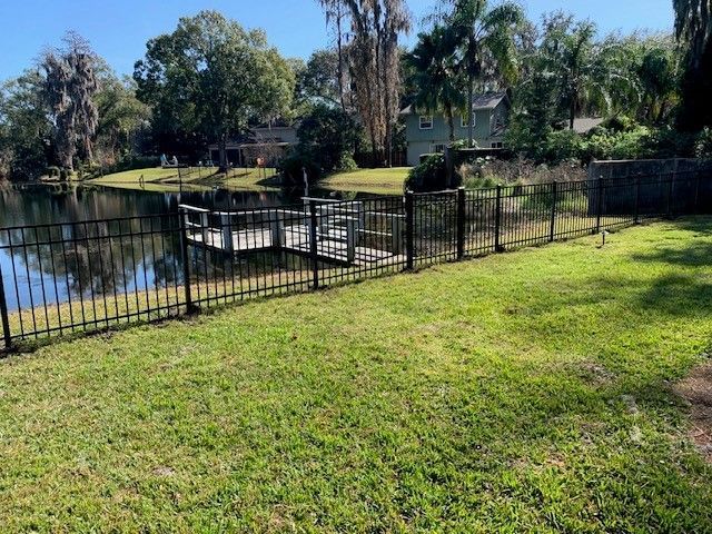 Lawn with black fence bordering a lake with a dock, trees in the background, sunny day.