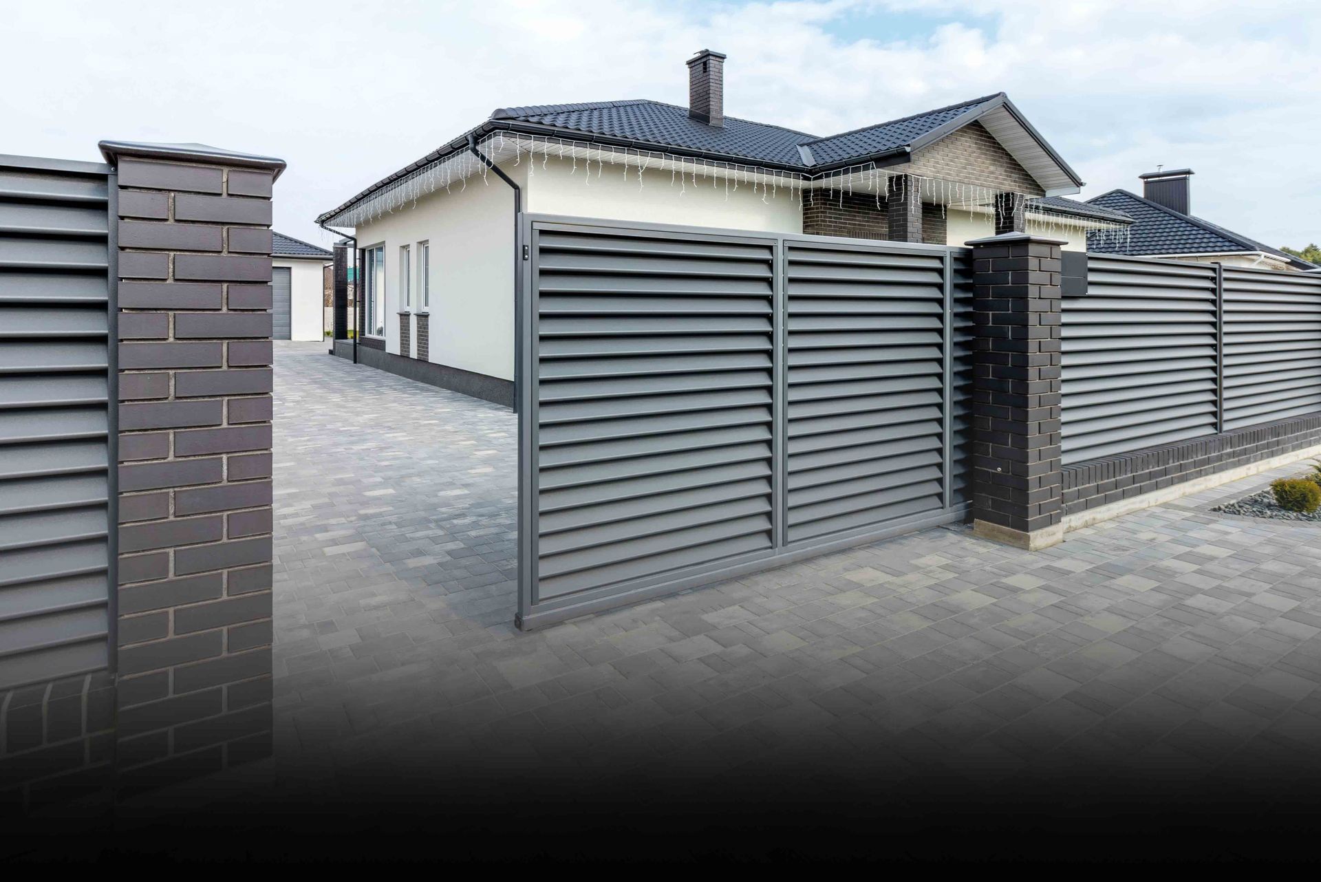 Modern house with gray horizontal slat fence and gate on brick pillars, paved driveway.