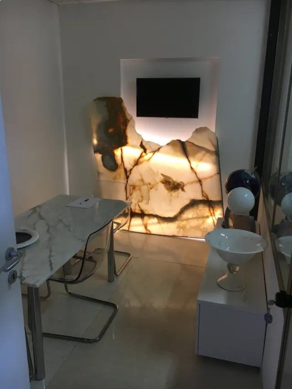 Piccola stanza con parete in pietra retroilluminata, TV, tavolo in marmo, mobile bianco e ciotola decorativa.