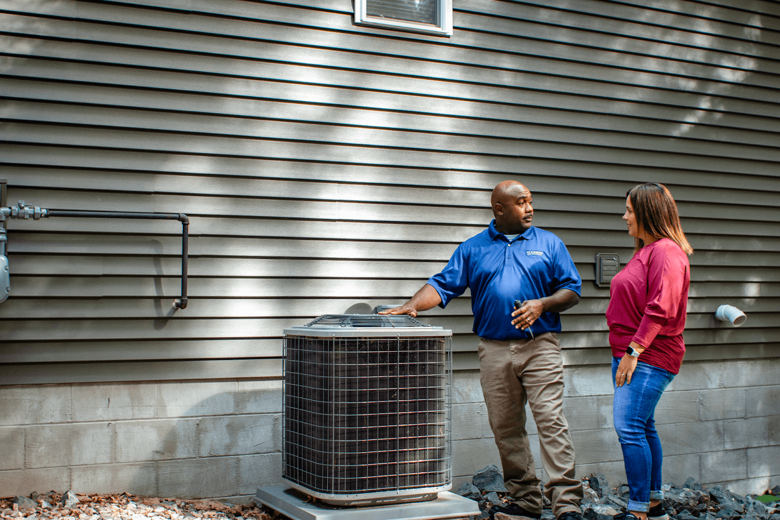 A man and a woman are standing in front of a house looking at an air conditioner.