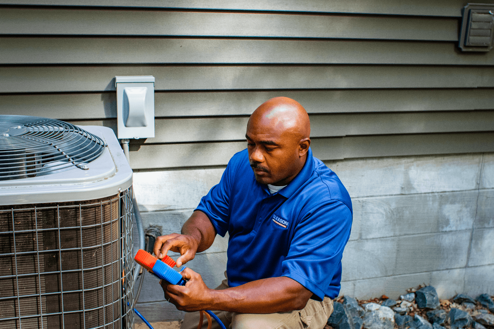 A man in a blue shirt is repairing on an air conditioner outside of a house.