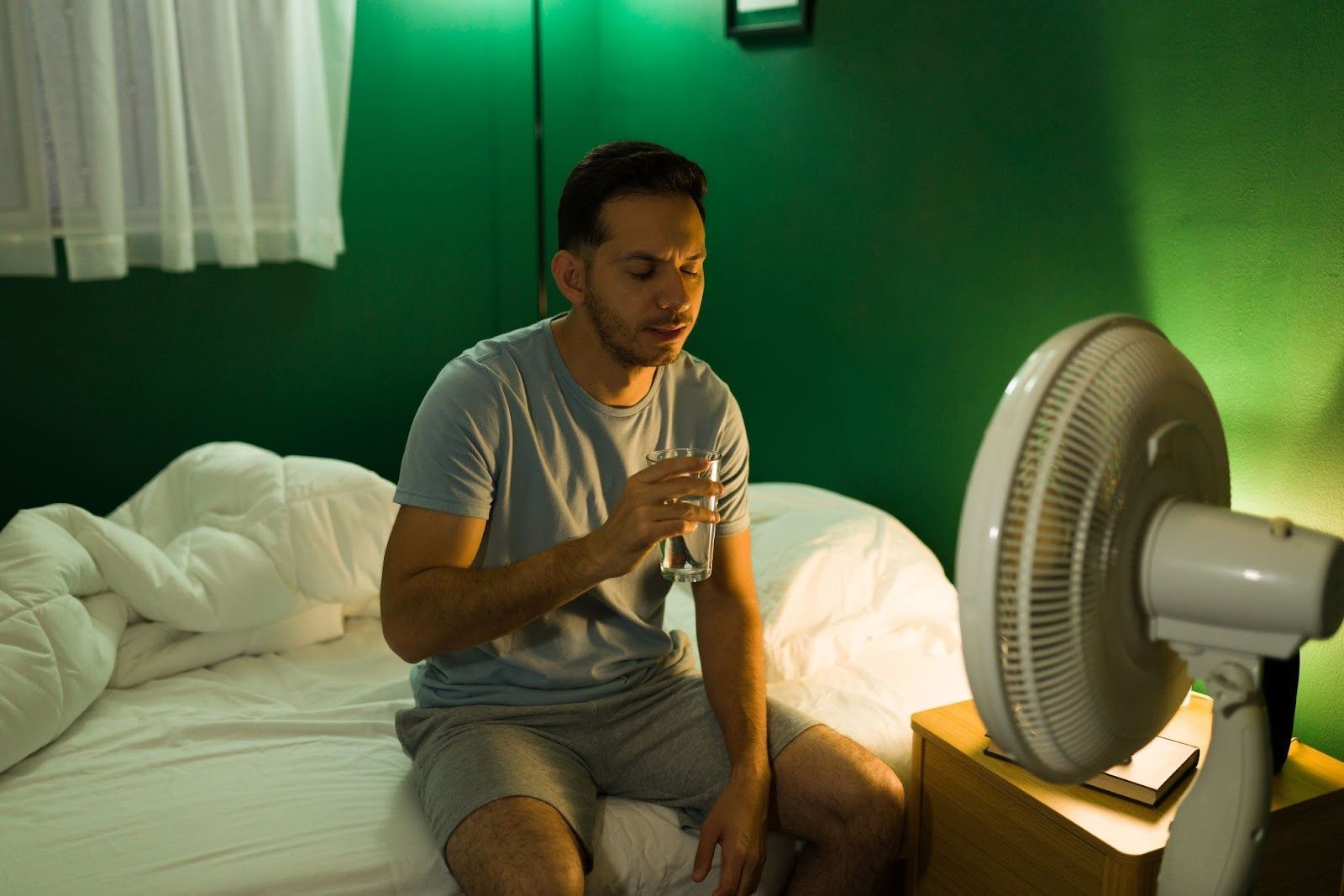 Man sits on side of bed in bright green room, drinking a glass of water and trying to cool off in front of table top fan 