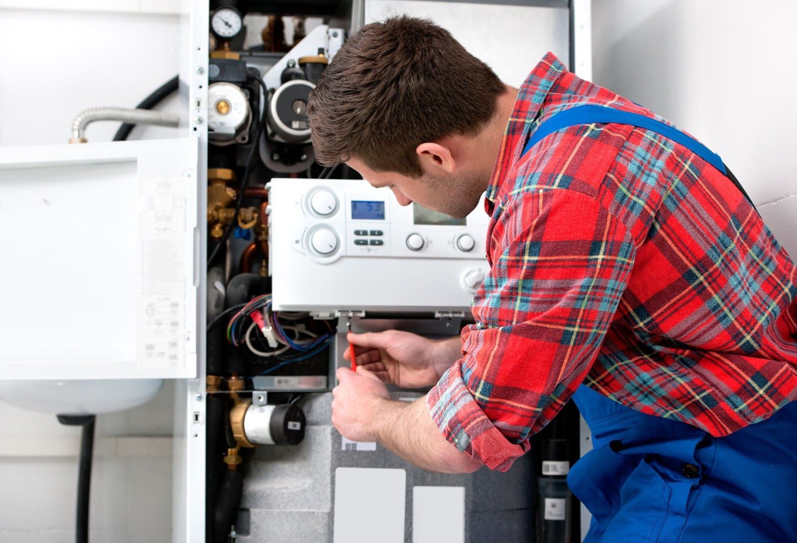 A man in a plaid shirt and blue overalls is working on a boiler.