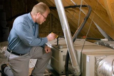 A man is kneeling down in an attic working on an air conditioner.