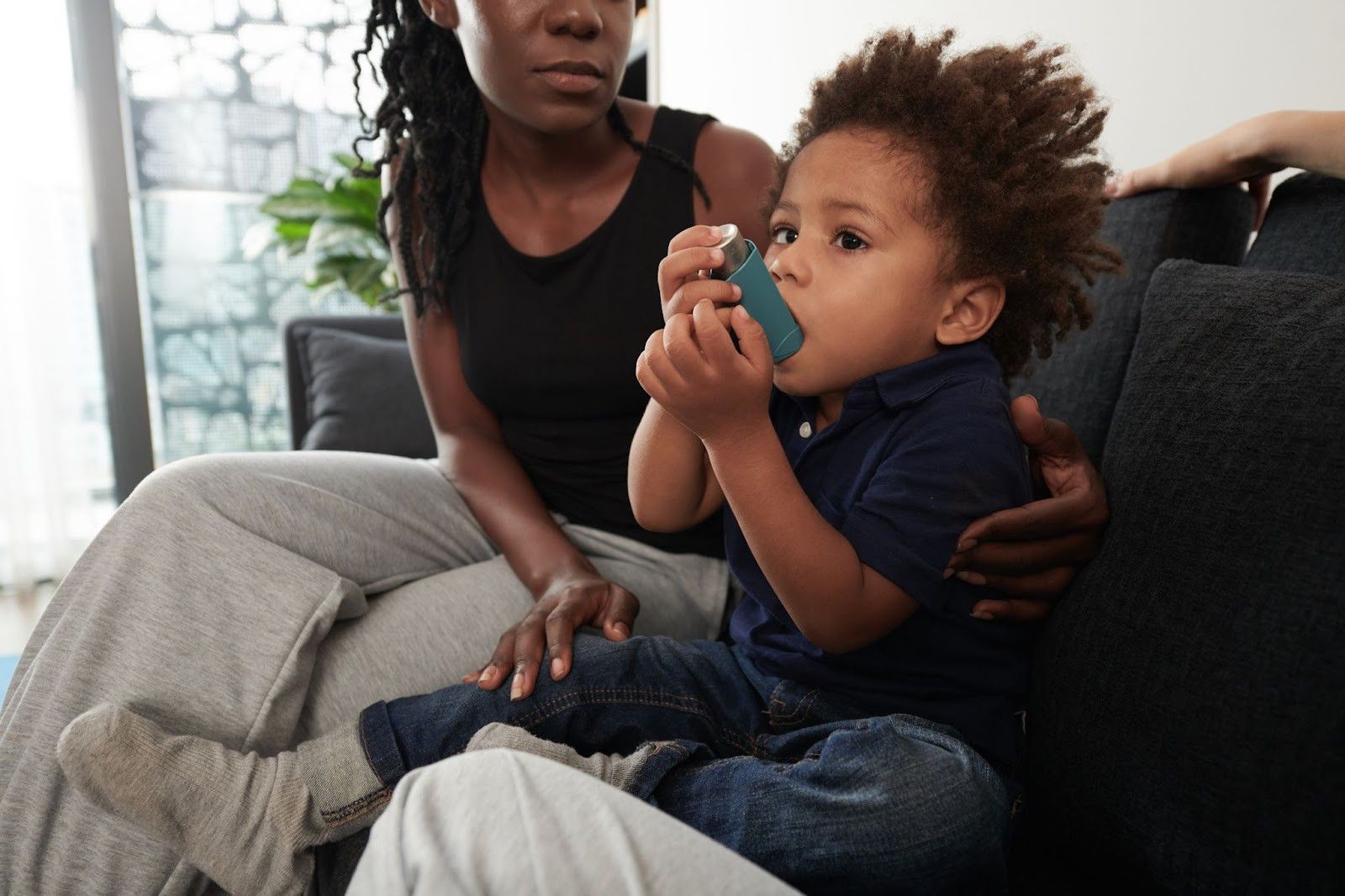 A woman is sitting on a couch with a child who is using an inhaler.